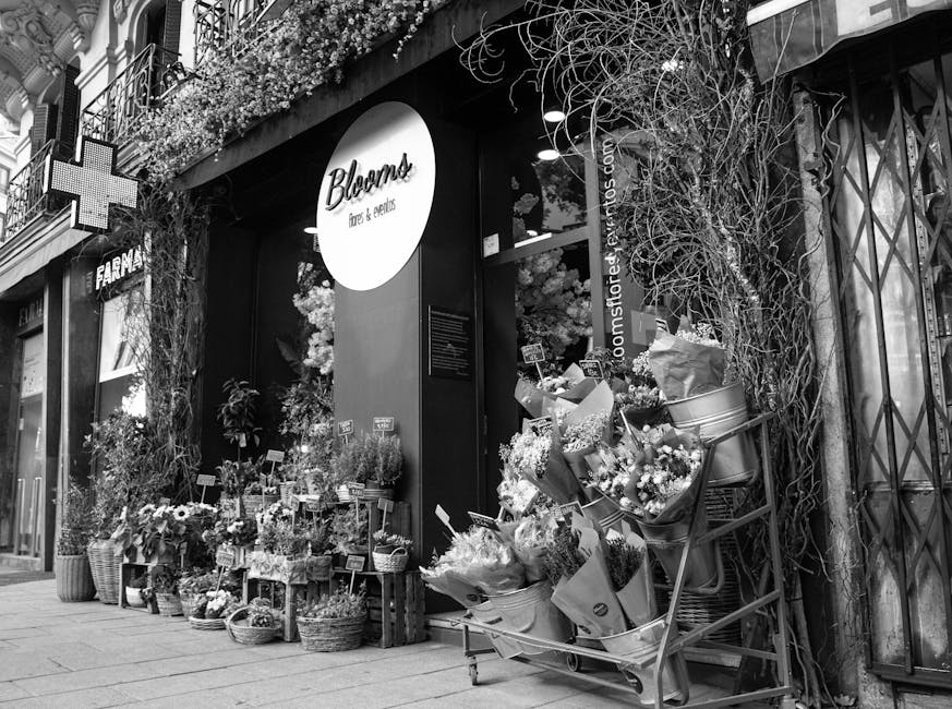 The storefront of Blooms, a flower shop on a city street, featuring a large circular sign with the shop's name. Outside, there is an array of fresh floral arrangements and bouquets in various wicker a