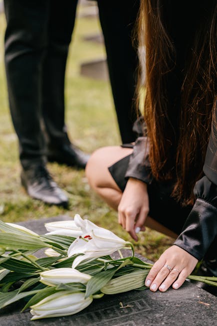 A person dressed in dark clothing kneels on the ground, arranging a floral tribute consisting of white lilies with large, smooth petals and green leaves. The bouquet is placed on a dark stone surface,