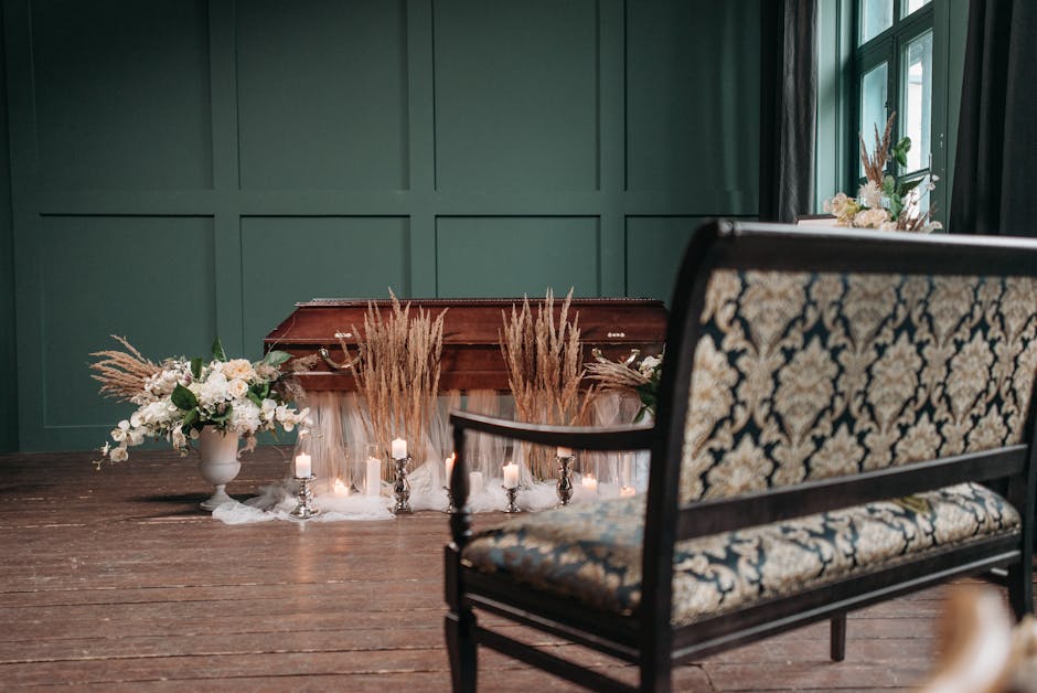 An arrangement of funeral flowers placed on a table in a room with dark green paneled walls and large windows. The floral display includes white roses, lilies, and other blooms in a white vase, comple