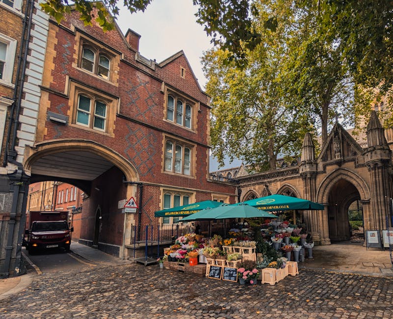 A colorful outdoor flower stand set up in front of a historic brick building with arched gateways in a cobblestone street. The stand is shaded by a green umbrella displaying the florist's website, and